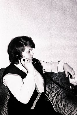 A woman sits on a patterned armchair, holding a vintage telephone headset. She wears a blouse and vest, suggesting a 1970s st...