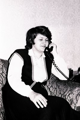 A woman sits on a patterned armchair, engaged in a phone call. She wears a dark, striped vest over a white blouse and a long,...