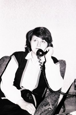 A woman seated, speaking into a vintage black rotary phone. She wears a white shirt, dark blazer, and striped vest. Mid-lengt...