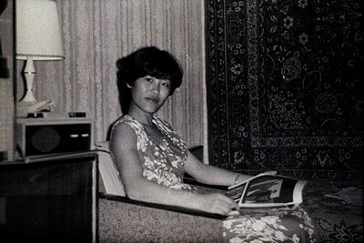Woman seated on a sofa, holding a magazine. Interior scene with a patterned rug, stereo, and table lamp. Appears to be a cand...