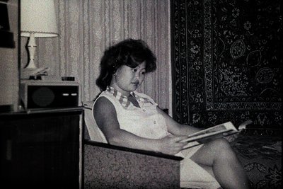 A woman seated in an armchair, engrossed in reading a book. The interior showcases mid-century modern design: a sleek lamp, v...