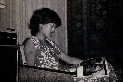 A woman seated in an upholstered chair, deeply engrossed in reading a magazine. She wears a floral-print dress, her hair styl...