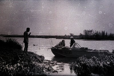 A small motorboat with the registration “KWS-6230” is docked near a shoreline, with three individuals present. One person sta...