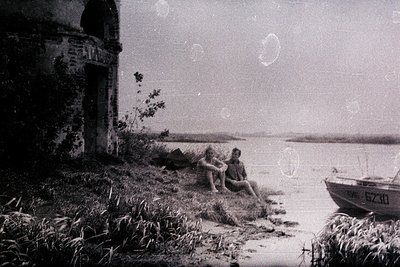 Two figures, a man and a woman, sit casually on grassy shoreline beside a small motorboat, with a crumbling stone building pa...