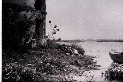 Overgrown, crumbling stone architecture frames a waterfront view. Two figures lie in tall grasses near the water’s edge, a sm...