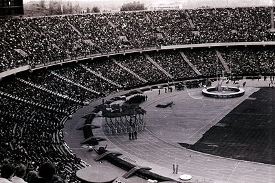 Massive, tiered stadium packed with spectators viewed from above. A ceremonial procession with flags & military personnel fea...