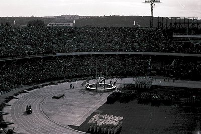 Panoramic view of a packed stadium during a ceremonial event. A motorcade enters the track, converging toward a central stage...