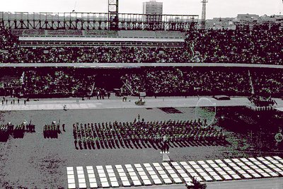 A large military parade takes place on a sprawling athletic field within a packed stadium. Rows of soldiers march in formatio...