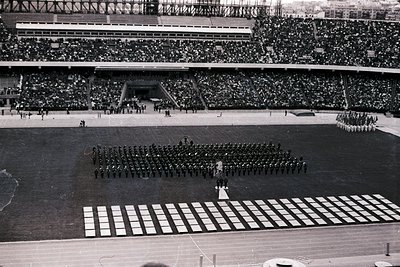 Massed military band and color guard perform on a track and field stadium. Tiered seating packed with spectators fills the ba...