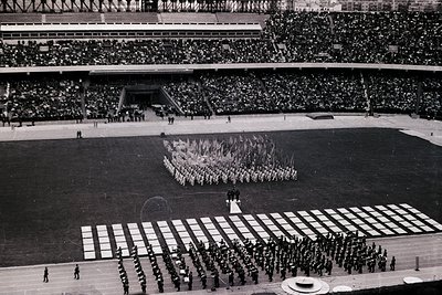 A wide, grayscale view captures a ceremonial parade formation on a large, grassy field within a massive stadium. Hundreds of ...