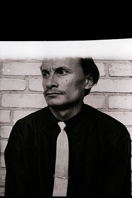 A black and white portrait of a man in a suit and tie against a textured brick wall. Strong studio lighting creates high cont...