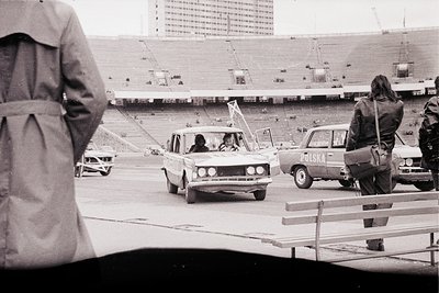 A partial view of a stadium, likely a sporting venue, dominates the background. Several vintage automobiles, including a Pols...