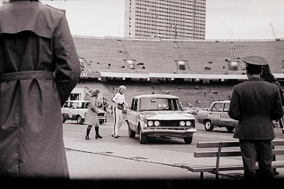 A street scene featuring a 1970s-era sedan and a woman in a long coat, likely in Eastern Europe. A uniformed officer observes...