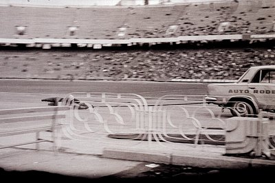 Vintage black and white photo shows a "Auto Rode" pickup truck parked beside a temporary barrier near a stadium. Packed stand...