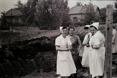 Four figures stand near a freshly dug area, possibly a construction site or garden. Two wear nurse’s caps and aprons, while t...