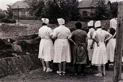 Group of women, likely nurses, stand observing a construction site or excavation. They are dressed in white uniforms and caps...