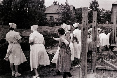 Group of women in medical attire and head coverings appear to be assisting with outdoor labor, possibly a community project. ...