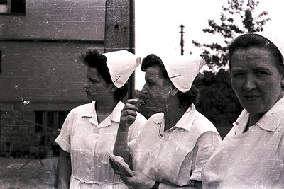 Three nurses in crisp white uniforms and distinctive caps stand outdoors, likely during a break. The composition reveals arch...