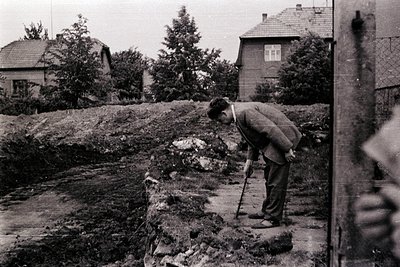 Man with cane leans forward, examining ground on a retaining wall overlooking a hillside & two houses. Likely a residential s...