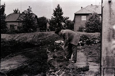 A man in a suit crouches, tending a small, overgrown garden on a brick ledge beside a modest home. The scene suggests a post-...