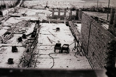 Construction site view, likely post-WWII. A brick wall under construction runs alongside excavated trenches. Visible equipmen...