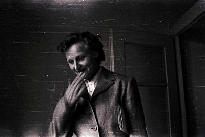 Portrait of an individual in a vintage suit, hands clasped near mouth, set against a plain wall. Style suggests mid-20th cent...