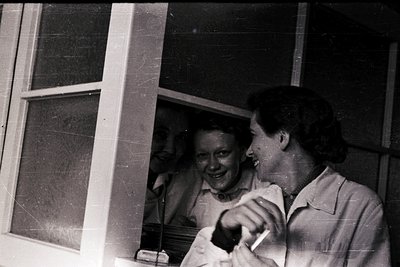 Two women in mid-20th-century lab coats pose indoors near a window, smiling at the camera. The setting appears clinical, like...