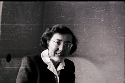 Mid-century woman in a sharp blazer and glasses, posed indoors against a plain wall. Classic 1950s–1960s fashion with rounded...