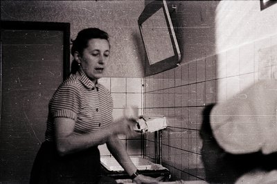 Mid-century bathroom scene with woman in striped blouse applying makeup at tiled sink. Mirror reflects partial view of anothe...