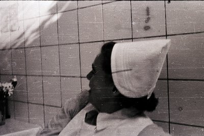 Vintage black-and-white portrait of a person in a large, conical headscarf, seated against a tiled wall. The setting appears ...