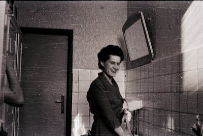 Vintage black-and-white photo of a woman in a hallway holding a rolled-up poster or banner. Mid-century tile walls and a door...