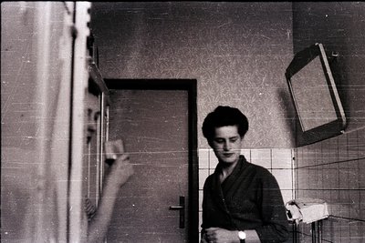 Vintage black-and-white photo of a young man in a tiled bathroom, holding a small wrapped package. Reflective mirror reveals ...