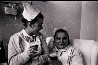 Two healthcare workers in 1950s-60s nursing uniforms—one in a cap—share a lighthearted moment indoors. The vintage black-and-...