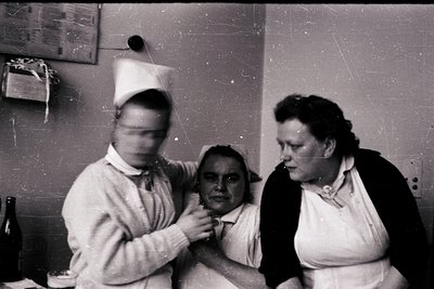 Mid-20th century medical scene: two nurses in white uniforms (one with a cap) attend to a seated patient in a clinical settin...