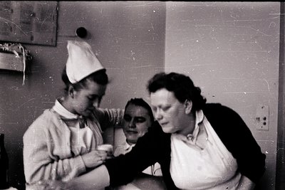 Two healthcare workers in 1950s-60s uniforms attend to a patient in a clinical setting. The nurse on the left wears a cap and...