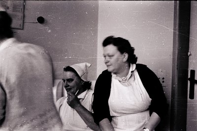 Mid-20th century black-and-white photo of two women in medical scrubs, likely nurses, in a clinical setting. One wears a cap;...