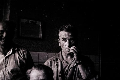 Black-and-white portrait of two men in a dimly lit interior, likely a 1950s–1960s setting. The man in focus wears a short-sle...