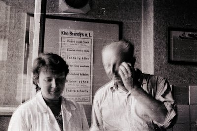 Two individuals in a vintage cinema setting, likely . The man adjusts the woman’s hair, while a poster for "Kino Brandys" lis...