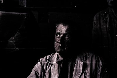 Portrait of an elderly man in low-light, likely mid-20th century. His weathered face and simple, high-collared shirt suggest ...