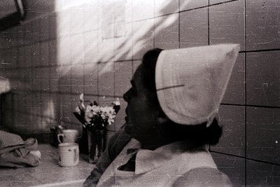 Vintage black-and-white photo of a woman in a nurse’s cap and uniform, seated in a tiled room with white tiles and a small wi...