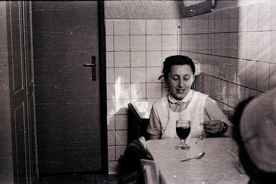 Mid-20th century indoor dining scene: woman in nurse’s uniform holds wineglass, seated at white-tiled table with blurred male...