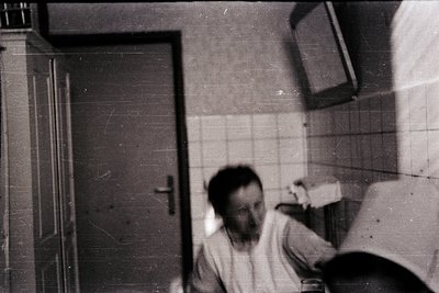 Vintage black-and-white photo of a man in a utilitarian bathroom with tiled walls and a closed door. His posture suggests he’...
