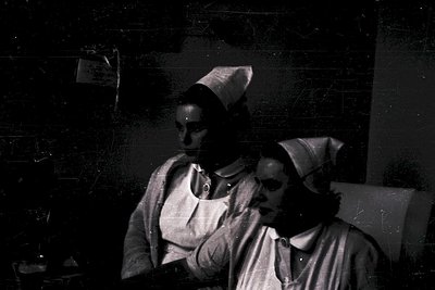 Vintage black-and-white photo of two women in nursing caps and uniforms, likely from a hospital or clinic setting. The woman ...