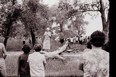 Black-and-white outdoor gathering in a wooded park setting, likely mid-20th century. A central figure in a patterned dress st...