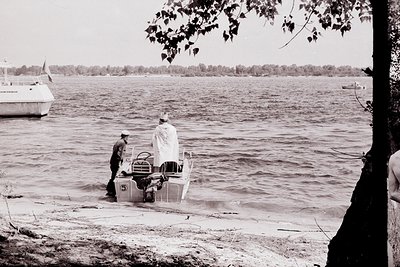 Black-and-white photo of two individuals assisting a small motorboat onto a sandy shore, likely mid-20th century. Boat featur...