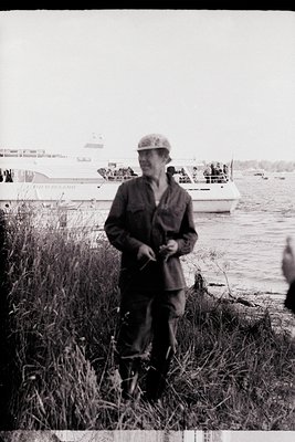 Mid-20th century black-and-white photo of a man in a military-style cap and workwear, standing by a waterfront with a fishing...