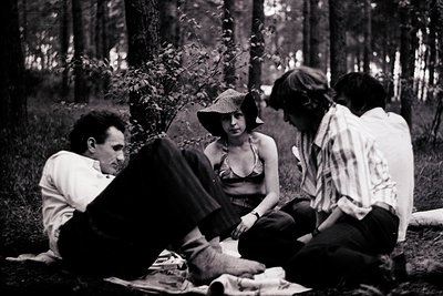 Four individuals seated on forest floor, mid-20th century. Man in white shirt kneels beside woman in wide-brimmed hat and bik...