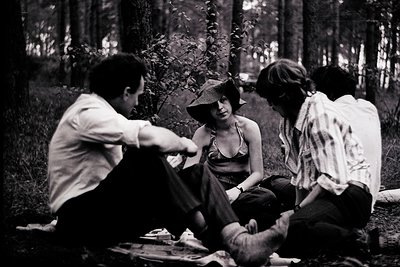 Four individuals seated in a wooded glade, mid-1970s. Man in light shirt leans forward, gesturing; woman in wide-brimmed hat ...