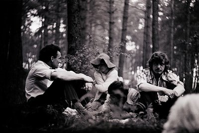 Black-and-white forest scene featuring three individuals seated on forest floor, likely mid-20th century. Dense pine trees fr...