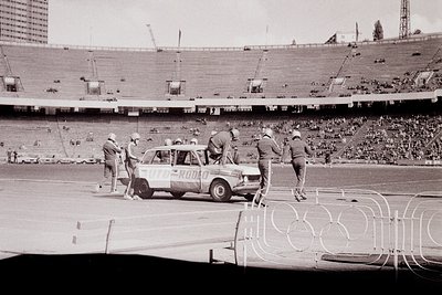 Black-and-white shot of a vintage Fiat 124 Sport Spider (1960s) being prepared on a stadium track during the **1968 Mexico Ci...